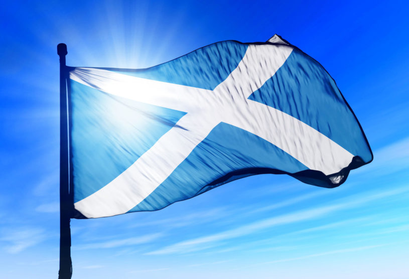 Scottish flag - blue with white x - waving in the wind with blue sky and sun in background.