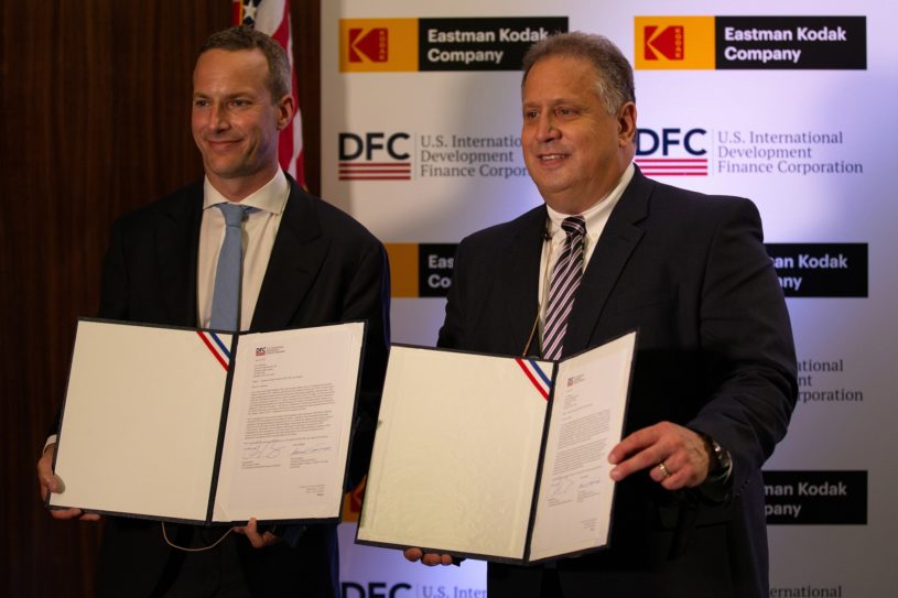 Two businessmen holding documents and showing them to a crowd to signify the receipt of a $765 million loan which caused a Kodak trading frenzy on Robinhood.