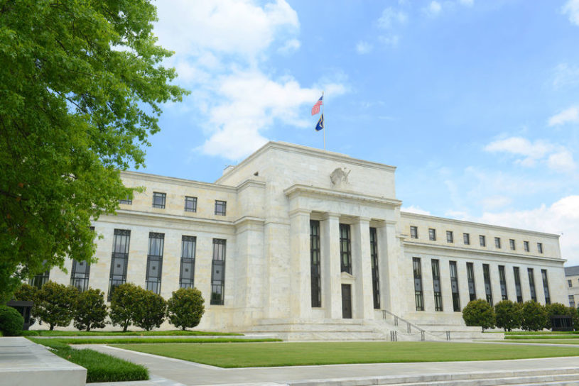 US Federal Reserve - large white, stone building with many pillars in front, blue sky in background, and US flag on top of building.