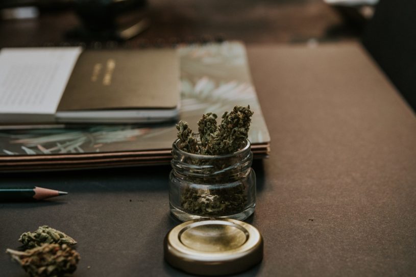 Jar of marijuana and paper stocks sitting on a wooden desk.