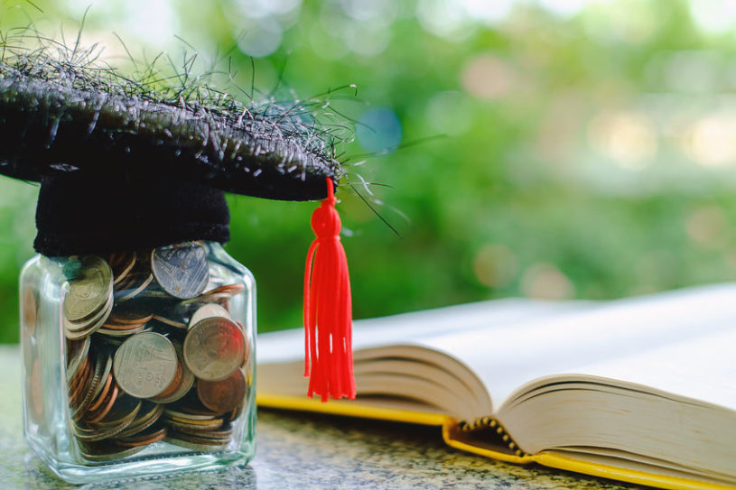 Graduation cap on a jar full of coins on a table, next to a yellow book about bitcoin