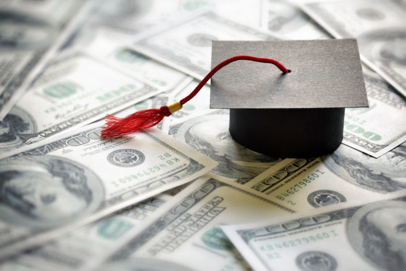 Several $100 USD notes spread out on a table with a black graduate cap