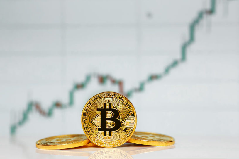 Several Bitcoin tokens in the foreground, stacked on each other, with a graph in the background showing the increased rate of Bitcoin adoption.