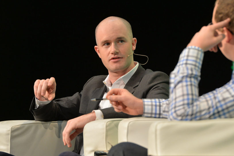 CEO of Coinbase wearing a business suit, sitting in a white chair, and talking to a crowd of millennials during a conference about Coinbase's new company culture.