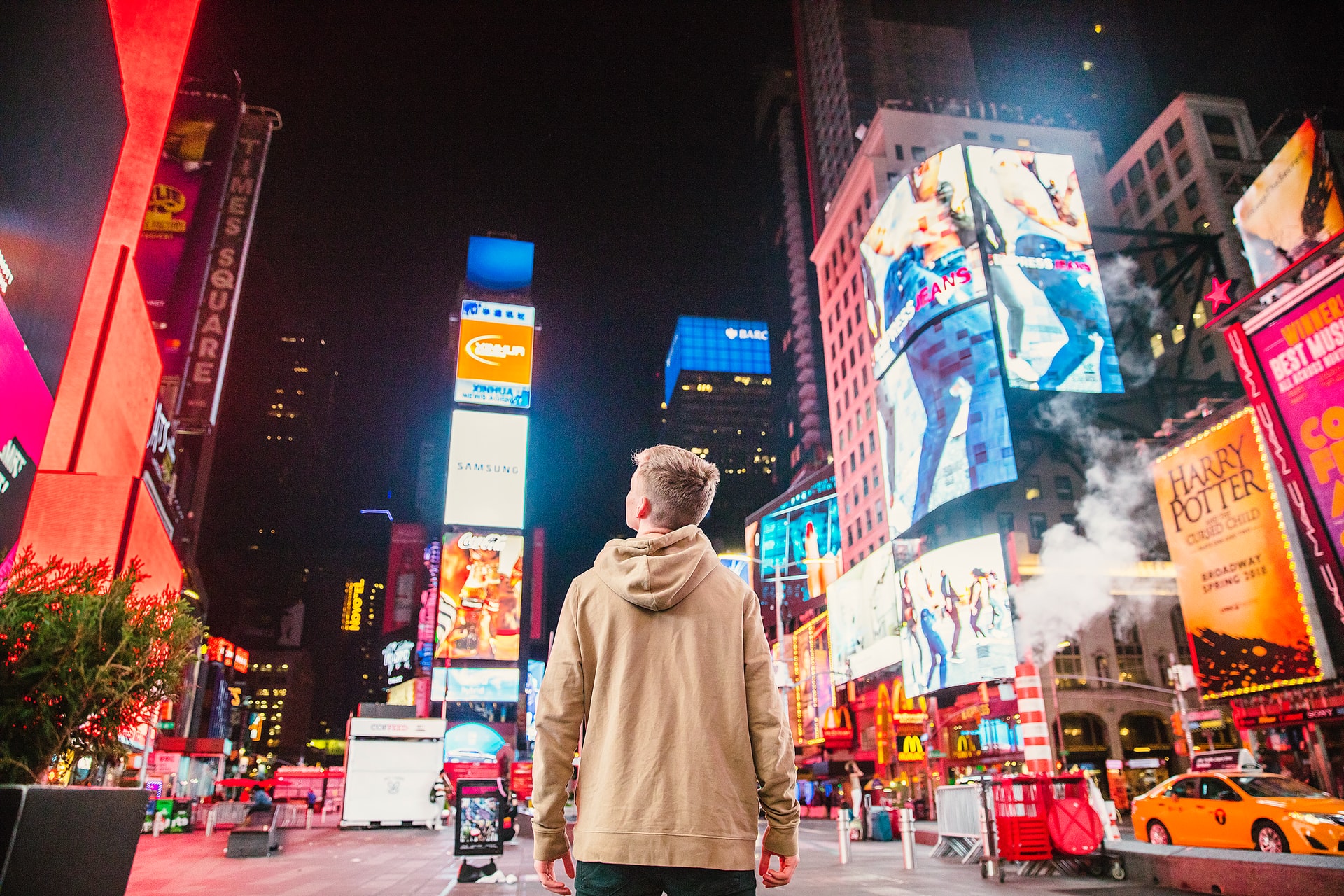 Young man with hooded sweatshirt standing in New York City looking up into the sky and lit buildings at night.