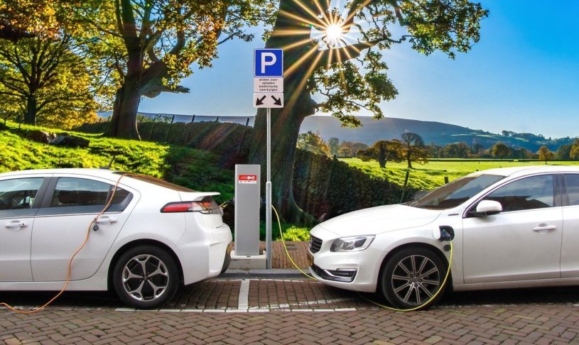 Two electric cars refeuling with sun and trees and blue sky in background.