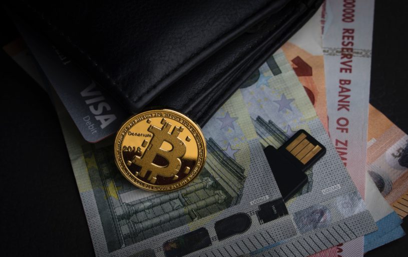 A coin, which represents Bitcoin is kept on top of a wallet, some currencies, and a YubiKey.