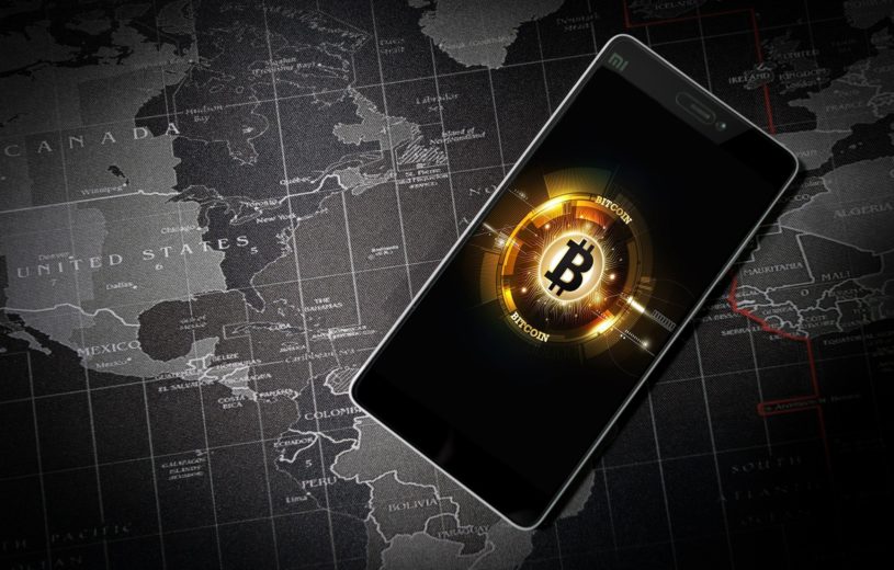 Bitcoin is shown in a mobile phone, which is placed over a world map.