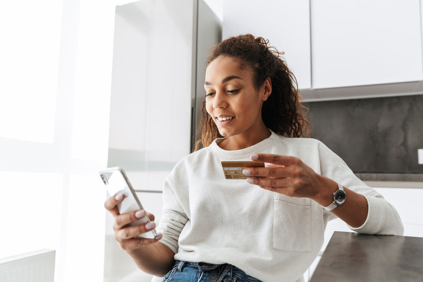 Image of young african american girl using mobile phone and credit card while sitting at table in bright living room