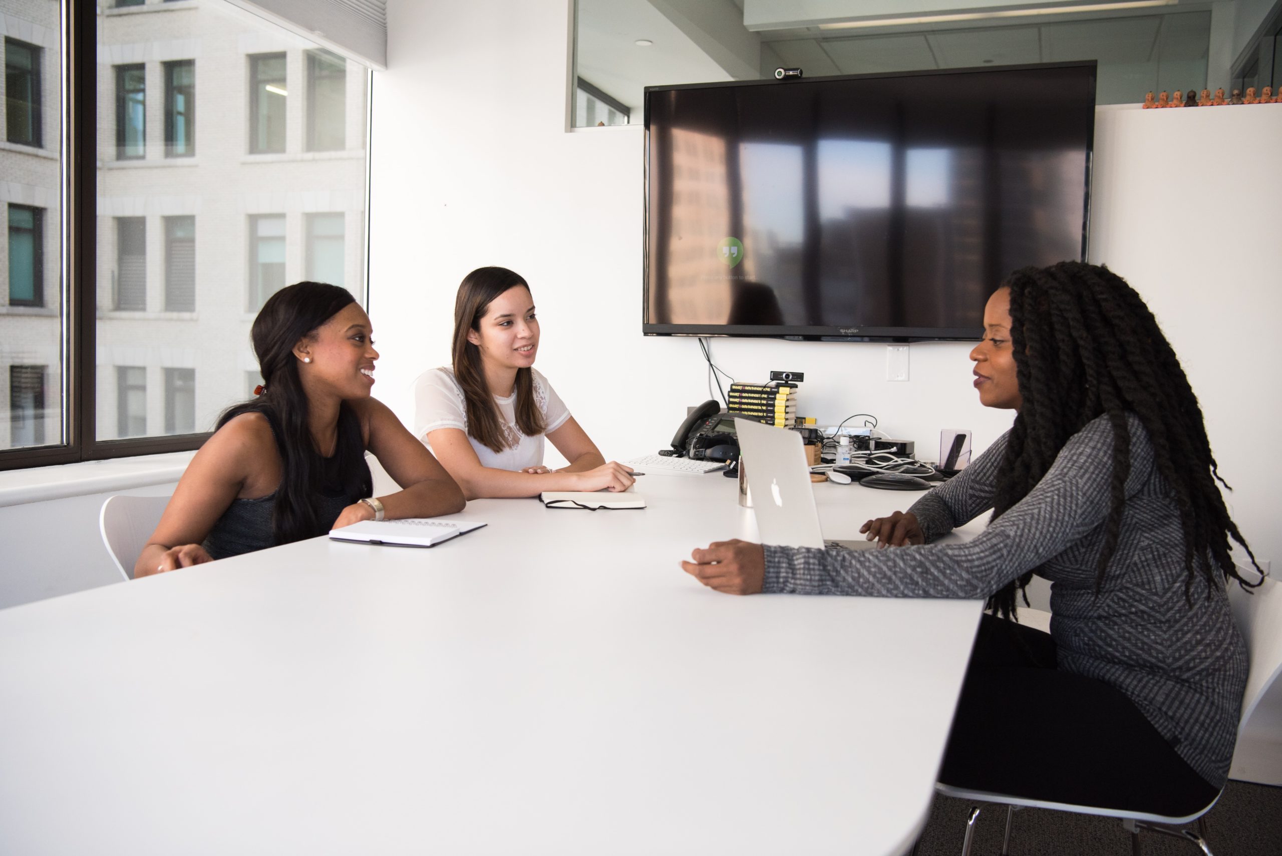 Three women sitting at a table in a conference room discussing trading cryptocurrencies on stock trading app Robinhood.