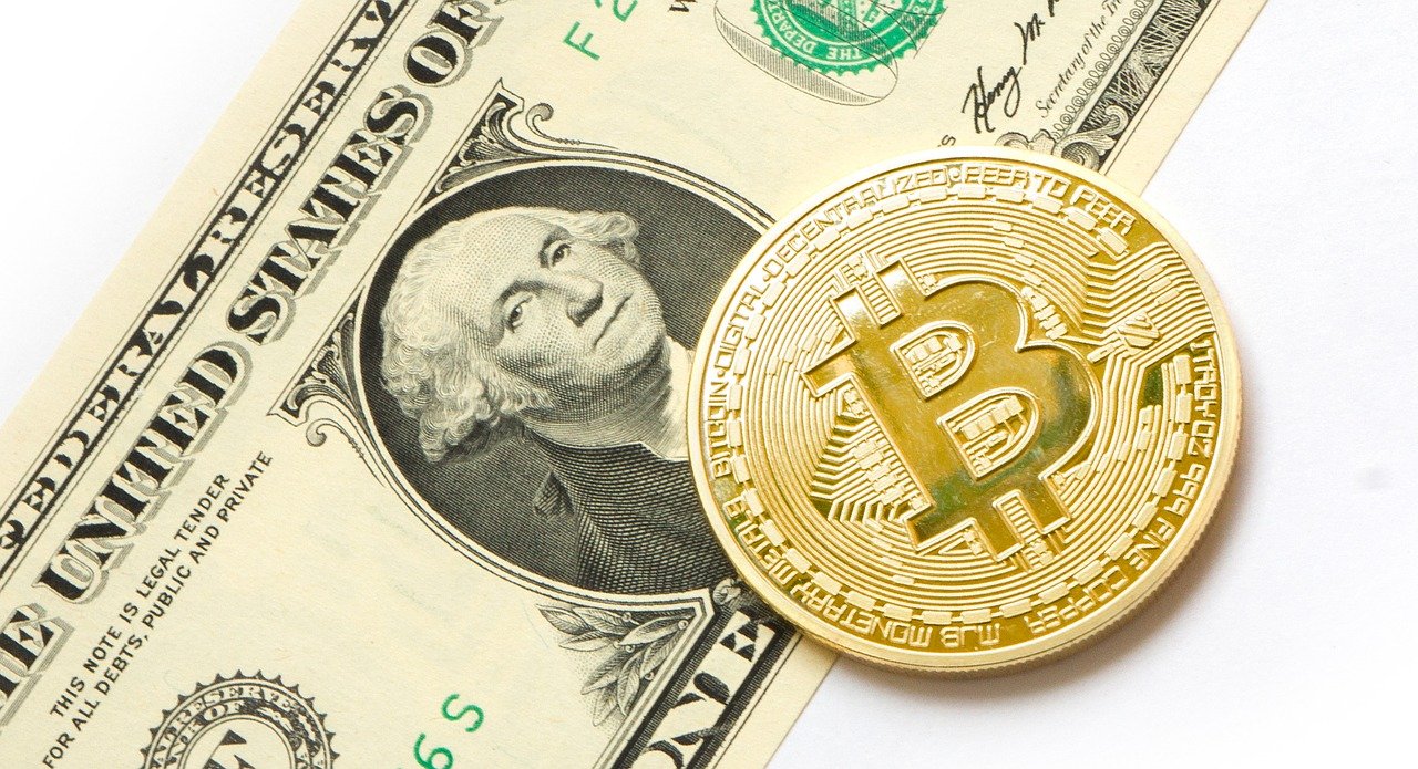 One Bitcoin token and one USD note laying on a white table.