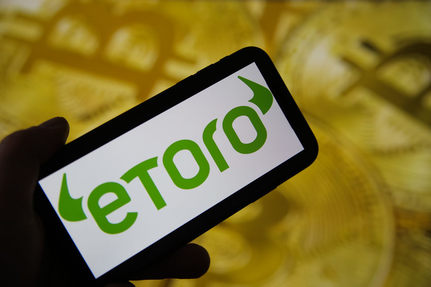 Closeup of mobile phone screen with logo lettering of etoro trading platform, blurred bitcoins background