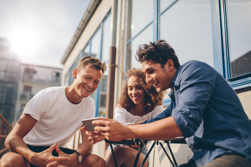 Three young friends sitting outdoors and looking at mobile phone. Group of people sitting at outdoor cafe and watching video on the smartphone, looking at altcoin gains.