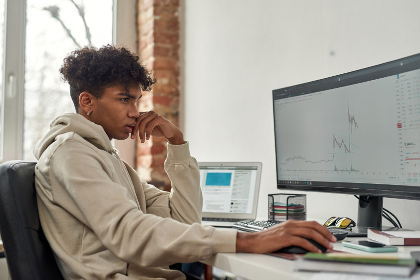 Side view of focused young guy trader looking at computer screen to analyze statistics while sitting at home office desk. Technology, stock market exchange, crypto currency