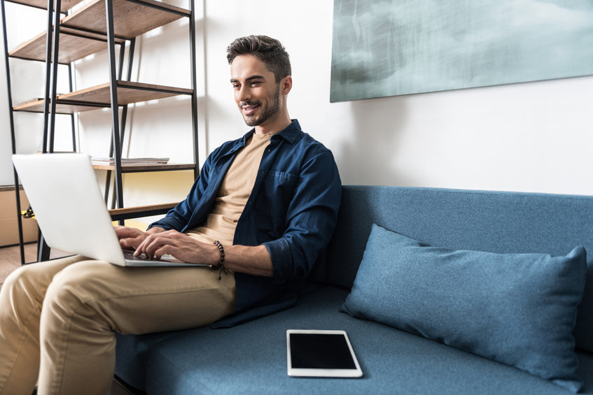 Cheery young bearded man is sitting on his home sofa and working with help of laptop computer. He is typing something and holding gadget on his laps. Copy space in right side