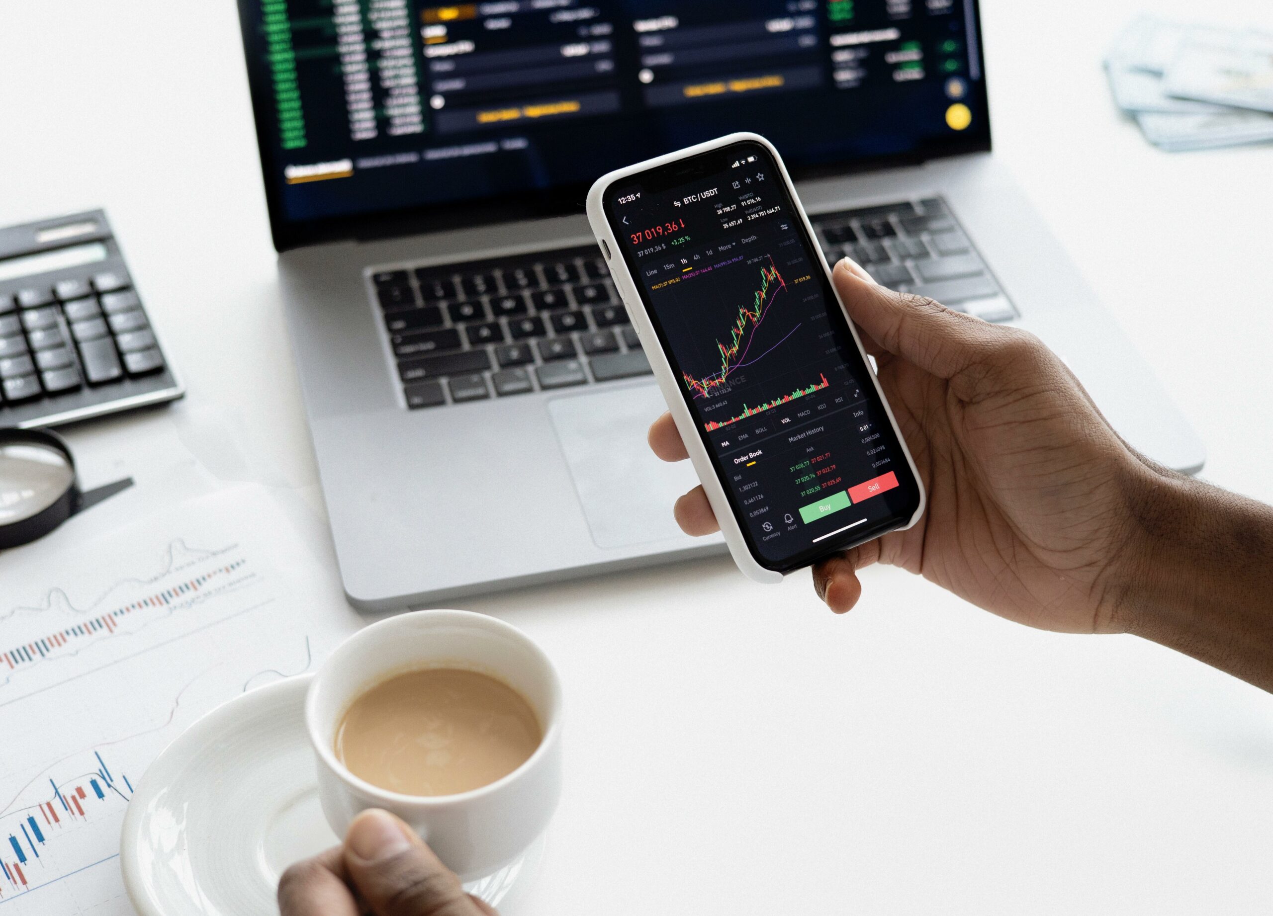 Someone at a desk on a mobile trading app with a cup of coffee