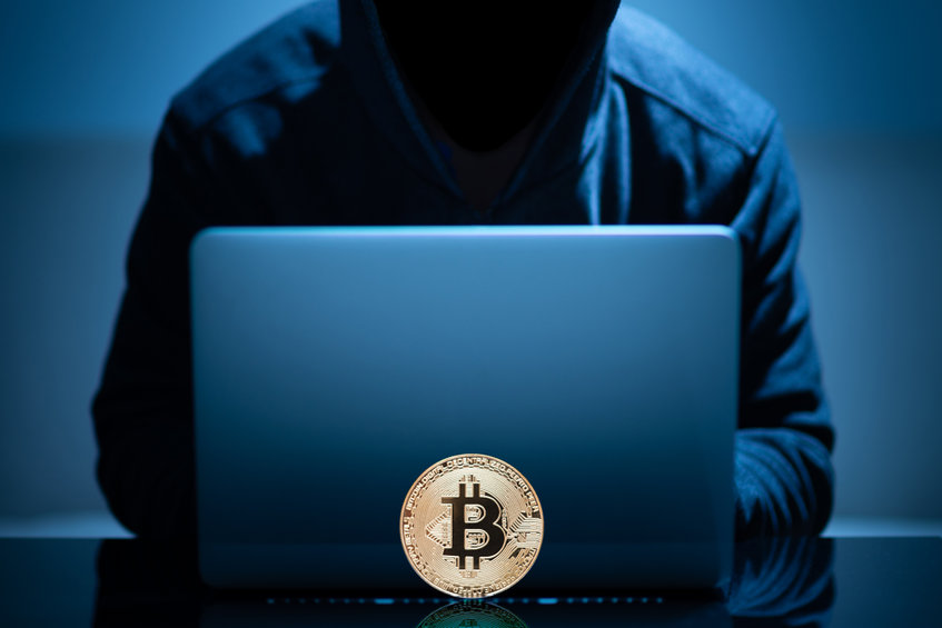 Image of hooded figure at laptop with a Bitcoin in foreground
