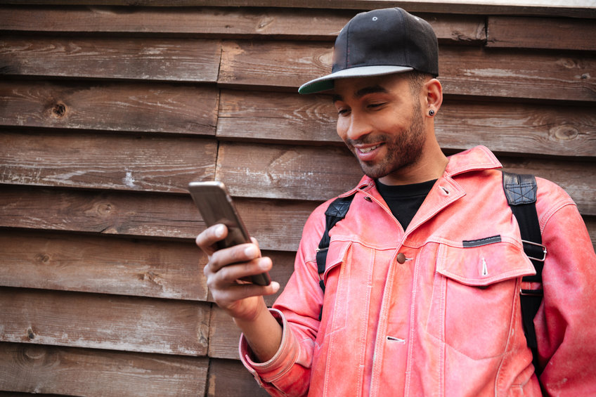Portrait of a happy smiling afro american man in hat typing message on mobile phone while standing outdoors on a street and reading about direct stock registration