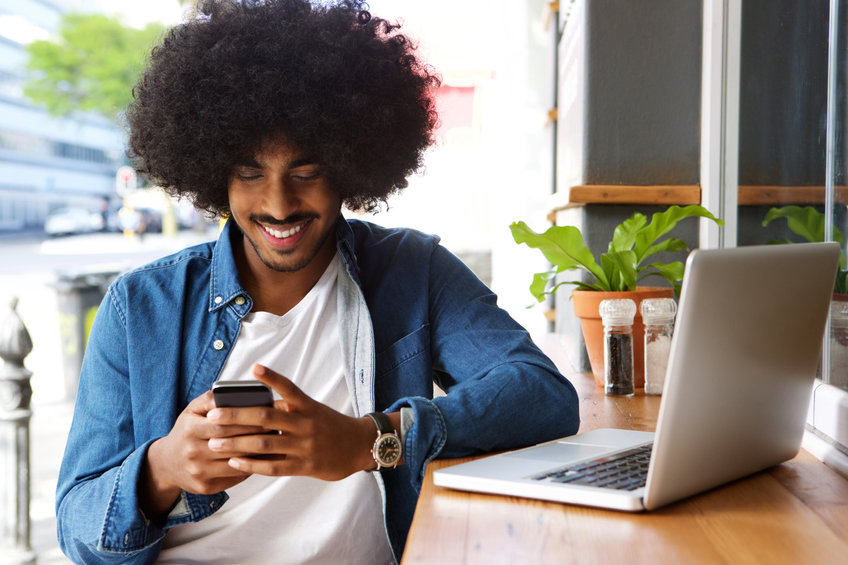 Portrait of a smiling guy using mobile phone and laptop to read about relationship between bitcoin and altcoins.