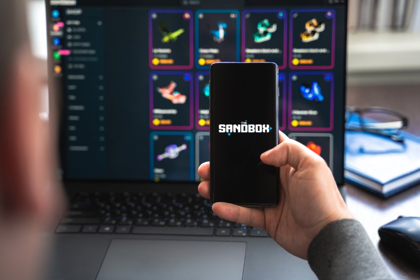 Image of a man holding a phone with The Sandbox's logo and a laptop in the background showing a NFT marketplace
