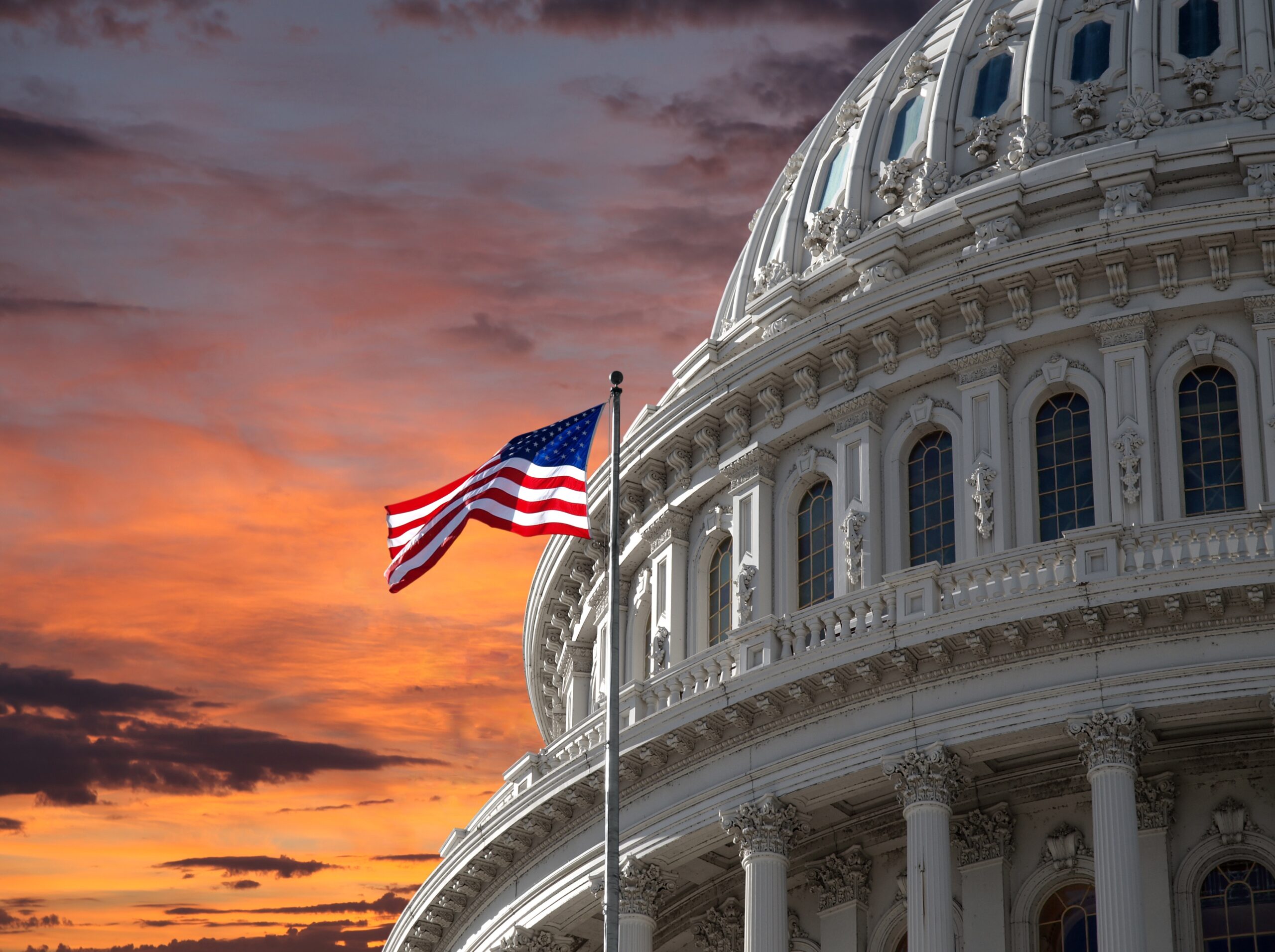 Dome of the US Capitol building with a US flag in front of the afternoon sky.