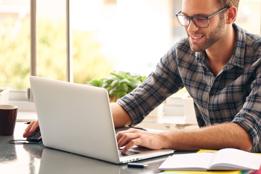 Happy young man, wearing glasses and smiling, as he works on his laptop