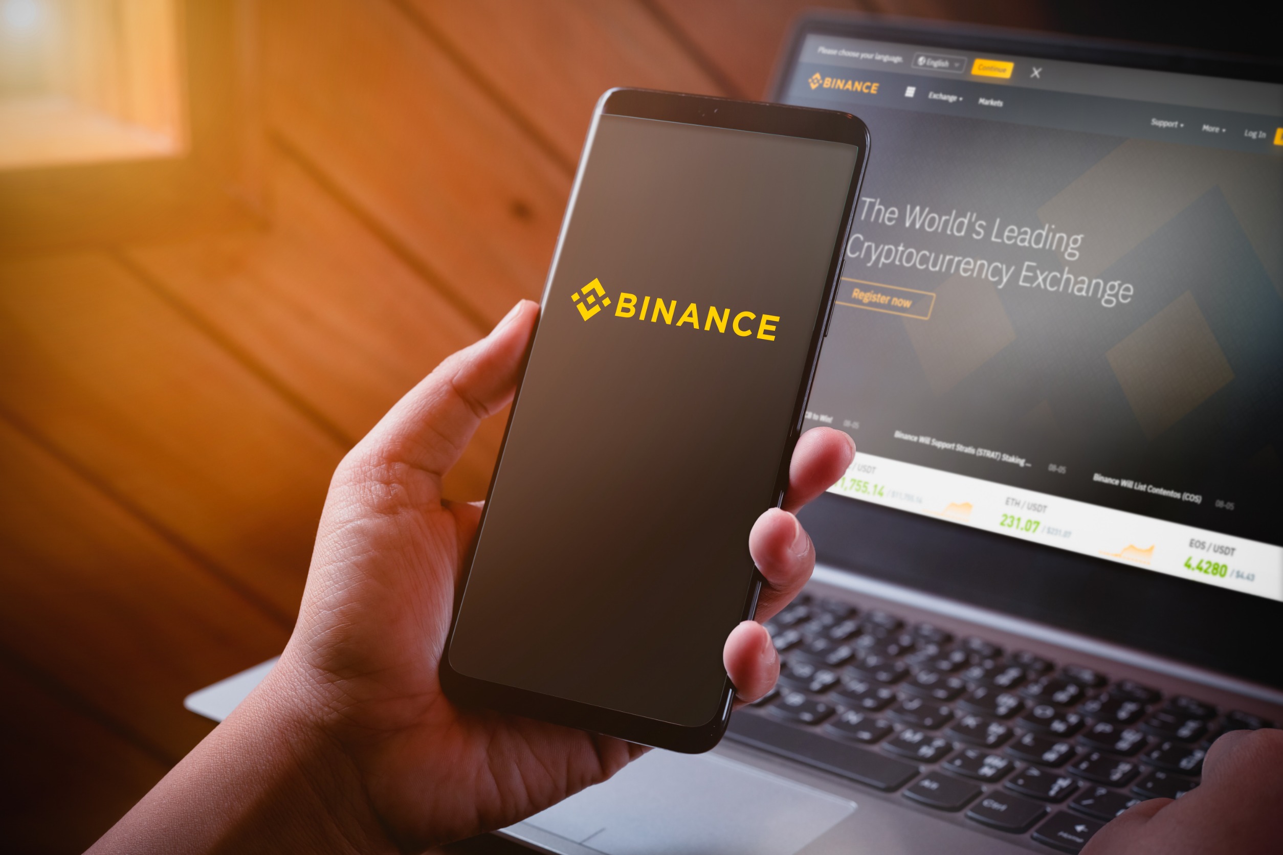 Hand holding a phone with "Binance" on screen in front of a laptop.