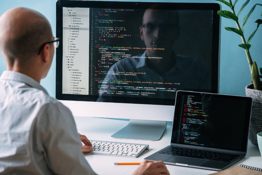 caucasian programmer in glasses is sitting behind the desk, in front of two black screens, laptop and monitor