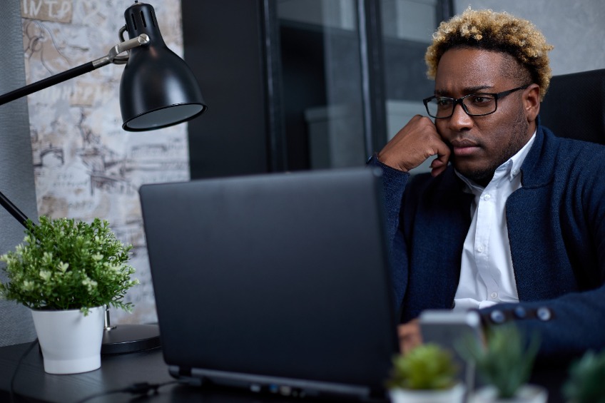 Thoughtful African man sitting at a table, working at a laptop, thinking about solving a problem, a thoughtful male employee thinking about an idea, looking at a computer screen, making a decision