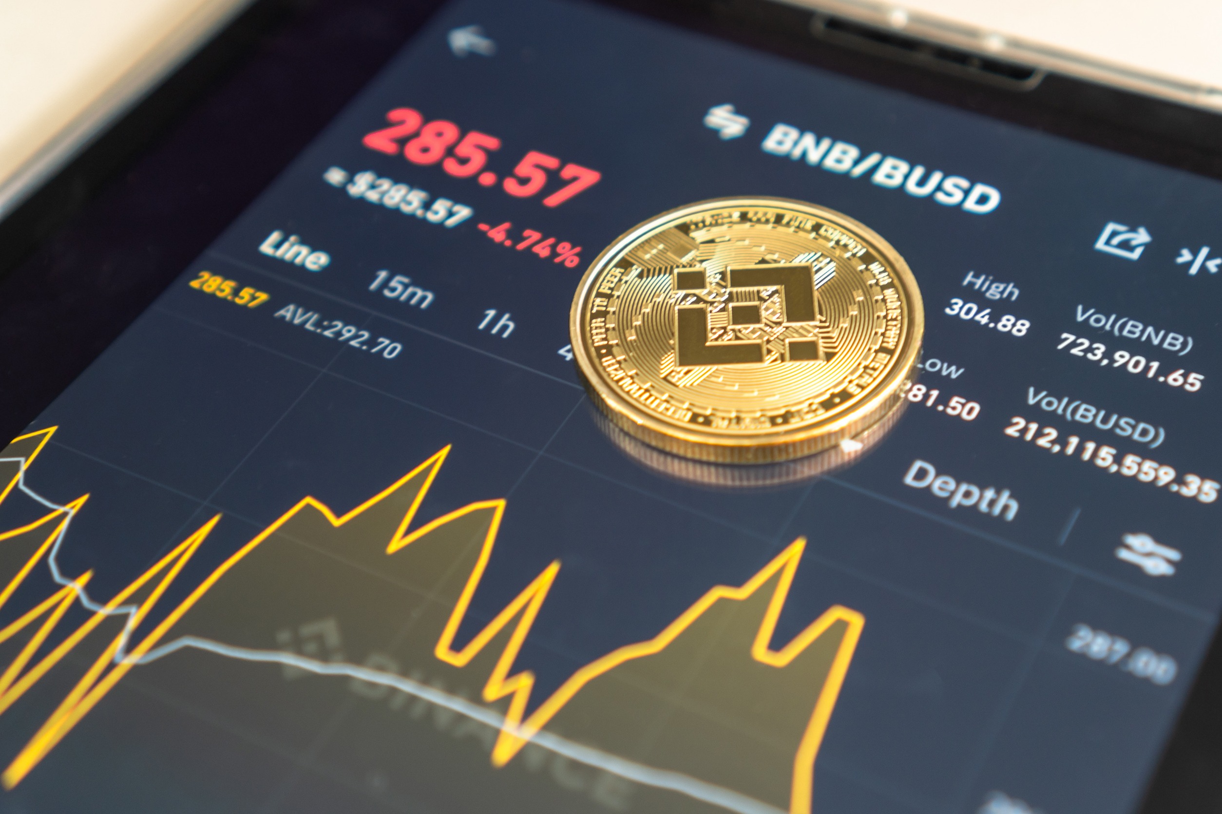 "BNB/BUSD" on a smartphone screen with a physical representation of the coin resting on top of the phone.