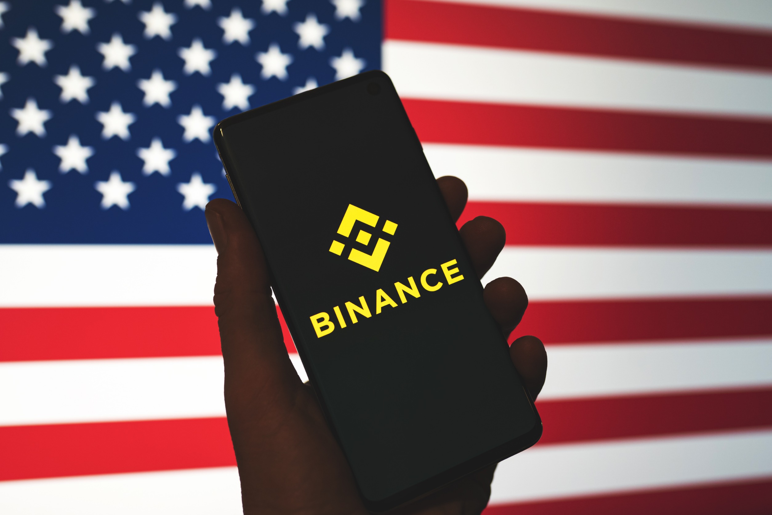 "Binance" on a phone screen in front of a large American flag
