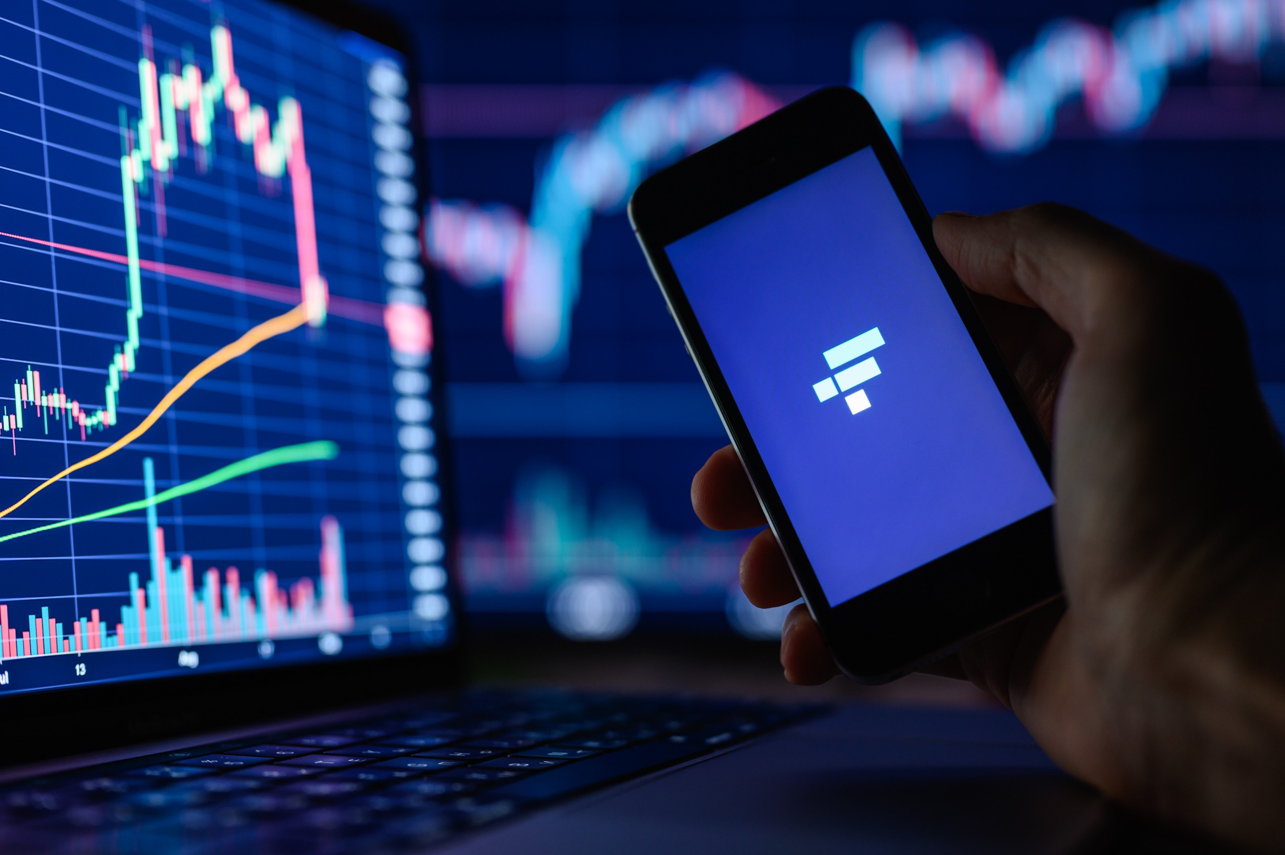 Smartphone showing FTX's "F" next to a compute showing a price chart which suddenly goes down after rising for some time.