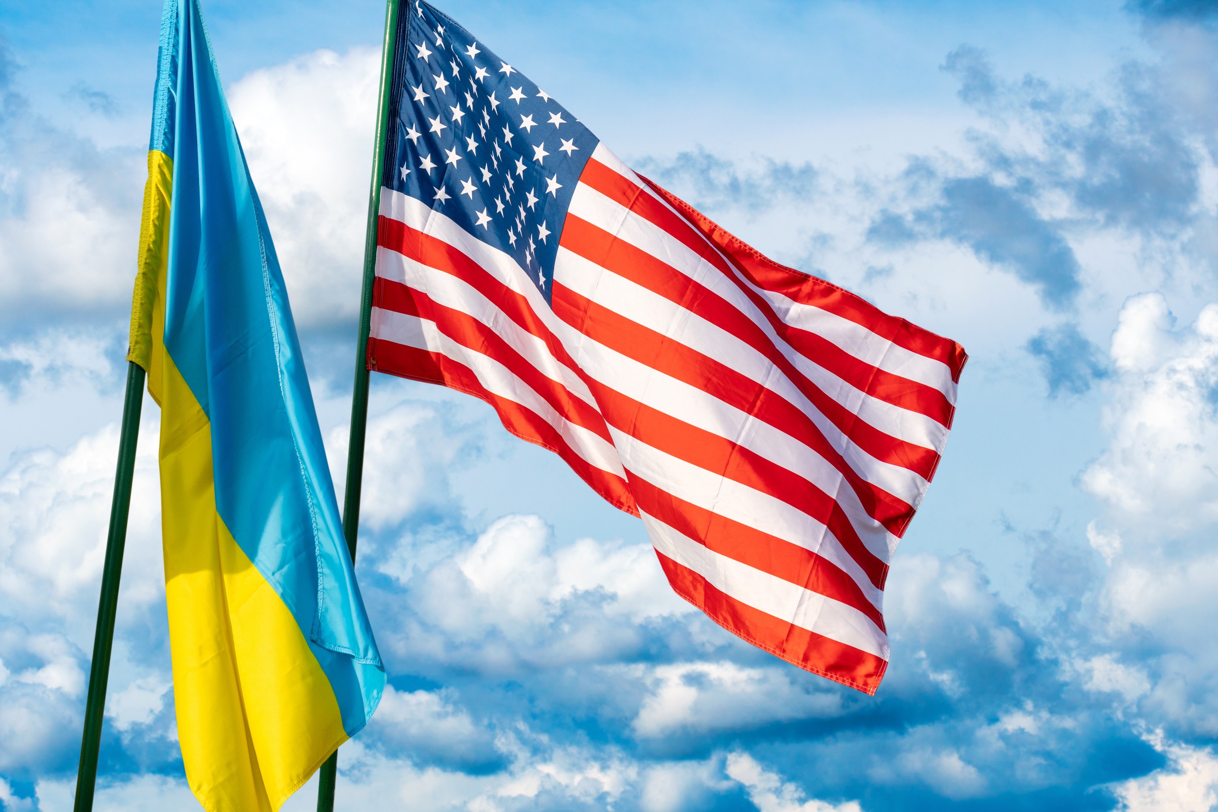Flags of USA and Ukraine next to each other on flagpoles against a blue sky.