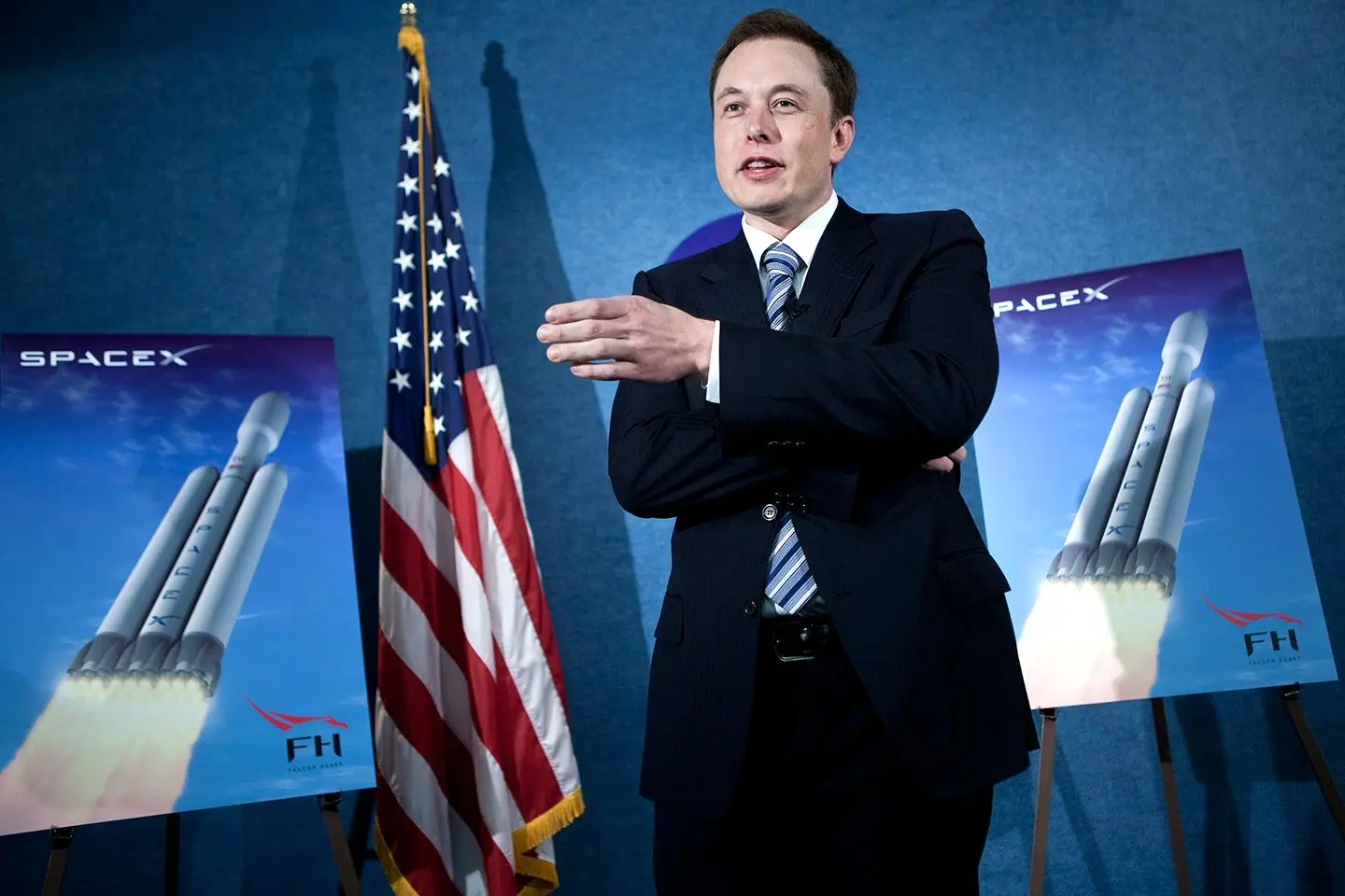 Elon Musk speaking alongside a SpaceX Falcon Heavy poster and American flag.